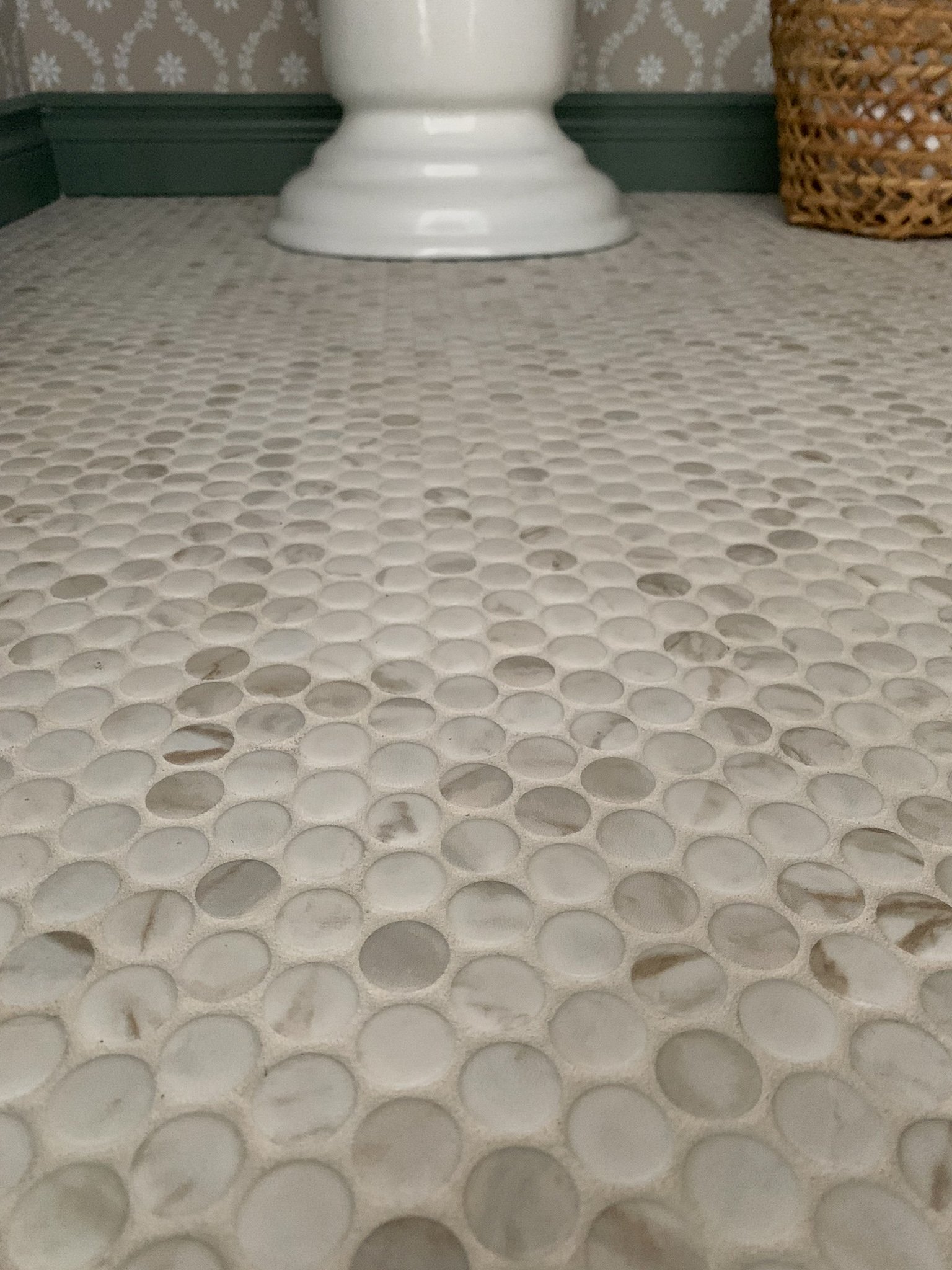 close up of marble look porcelain penny tile, with green trim and pedestal sink base and wicker basket in the background