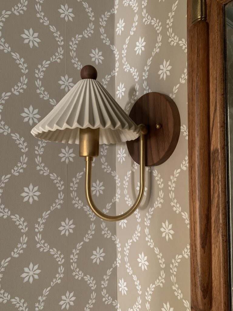 sconce with walnut wood base, brass arm and ceramic pleated shade on a floral wallpaper background