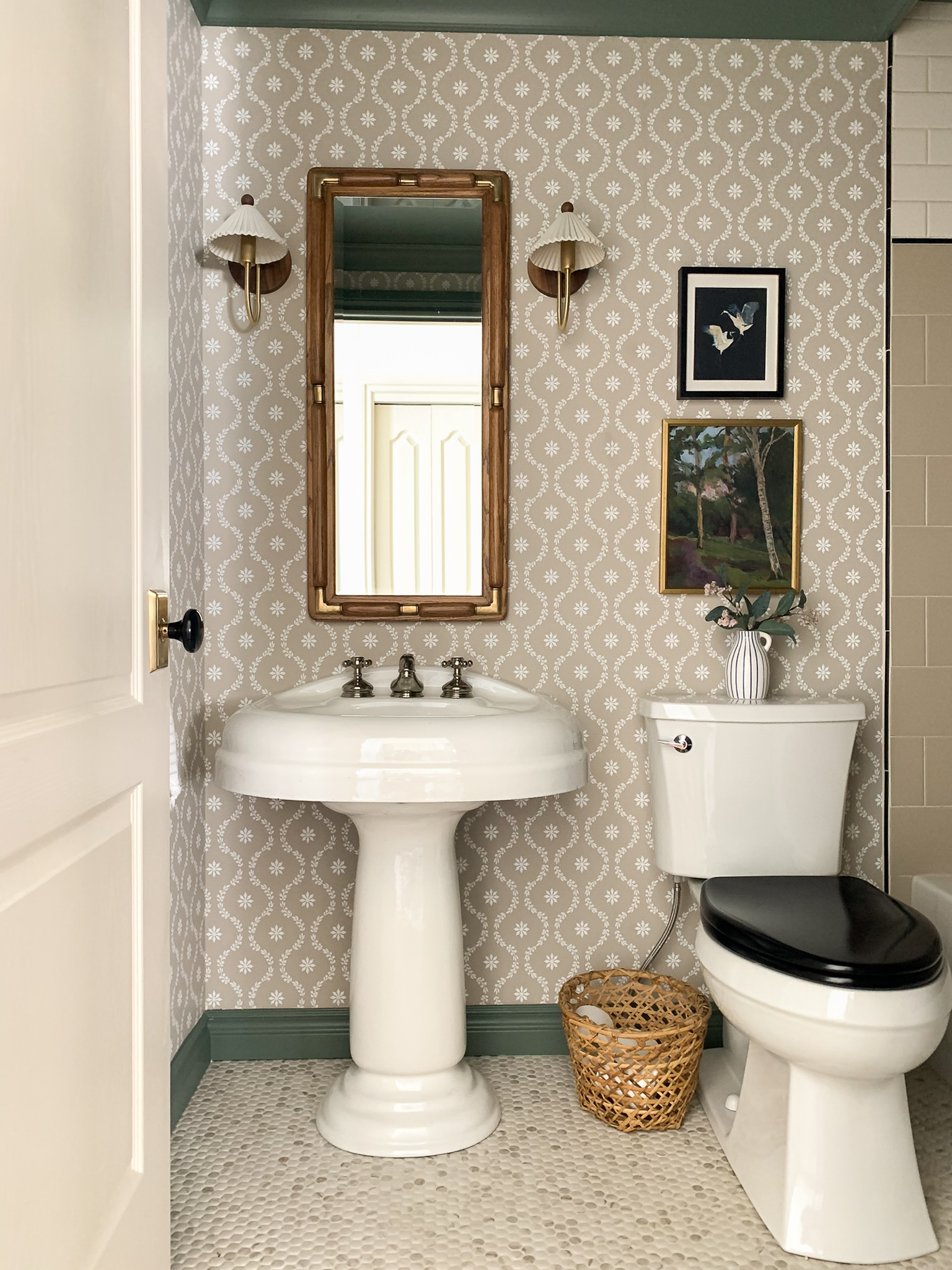 Modern tradtional bathroom with vintage pedestal sink, beige floral trellis wallpaper, penny tile floor, contrasting green trim and ceiling, toilet with a black seat.