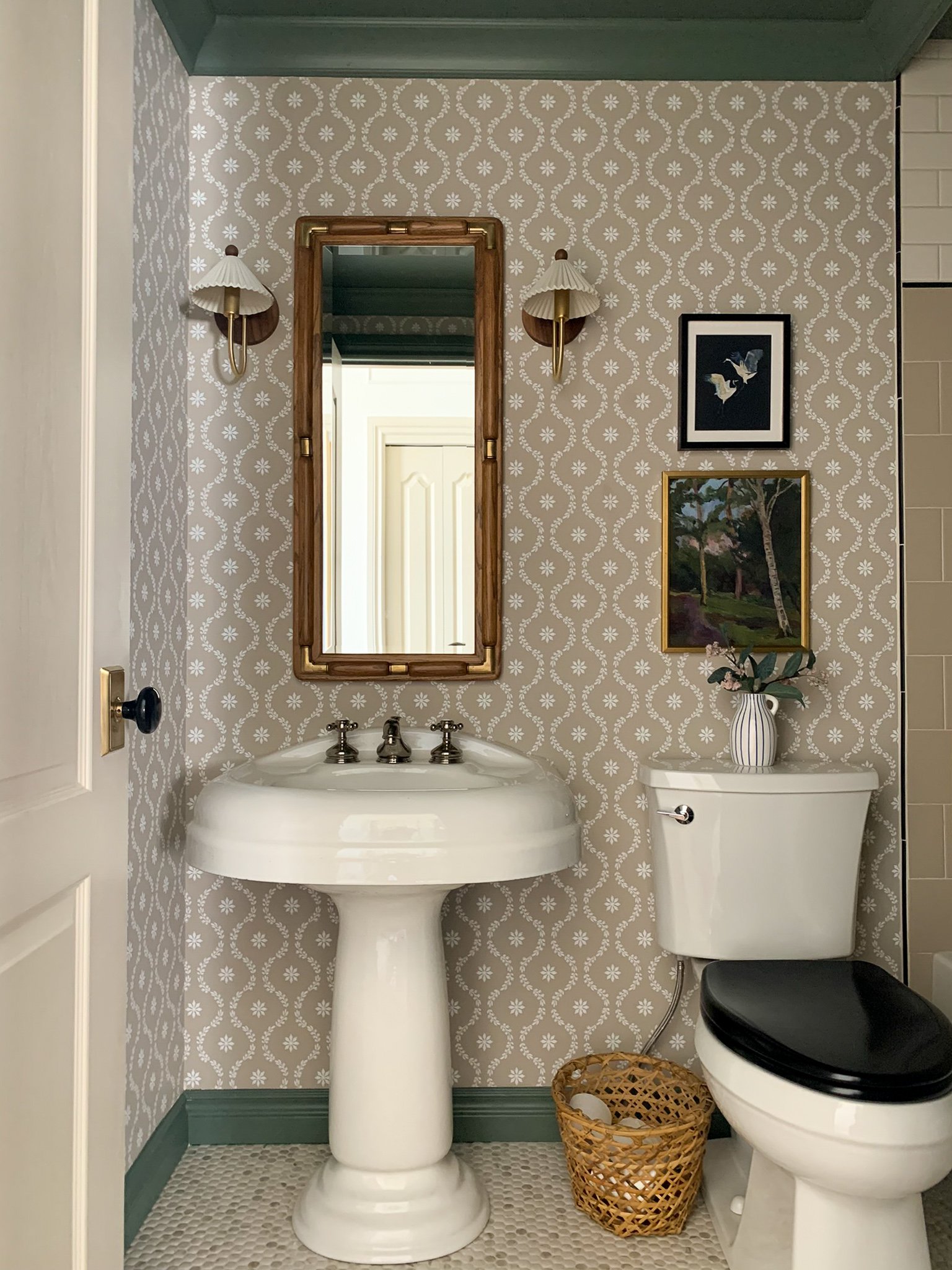 Modern traditional Bathroom with green ceiling and crown moulding, floral beige and white wallpaper, brass  and walnut scalloped shade sconces, vintage wood mirror, art stacked over toilet, penny tile and vintage pedestal sink