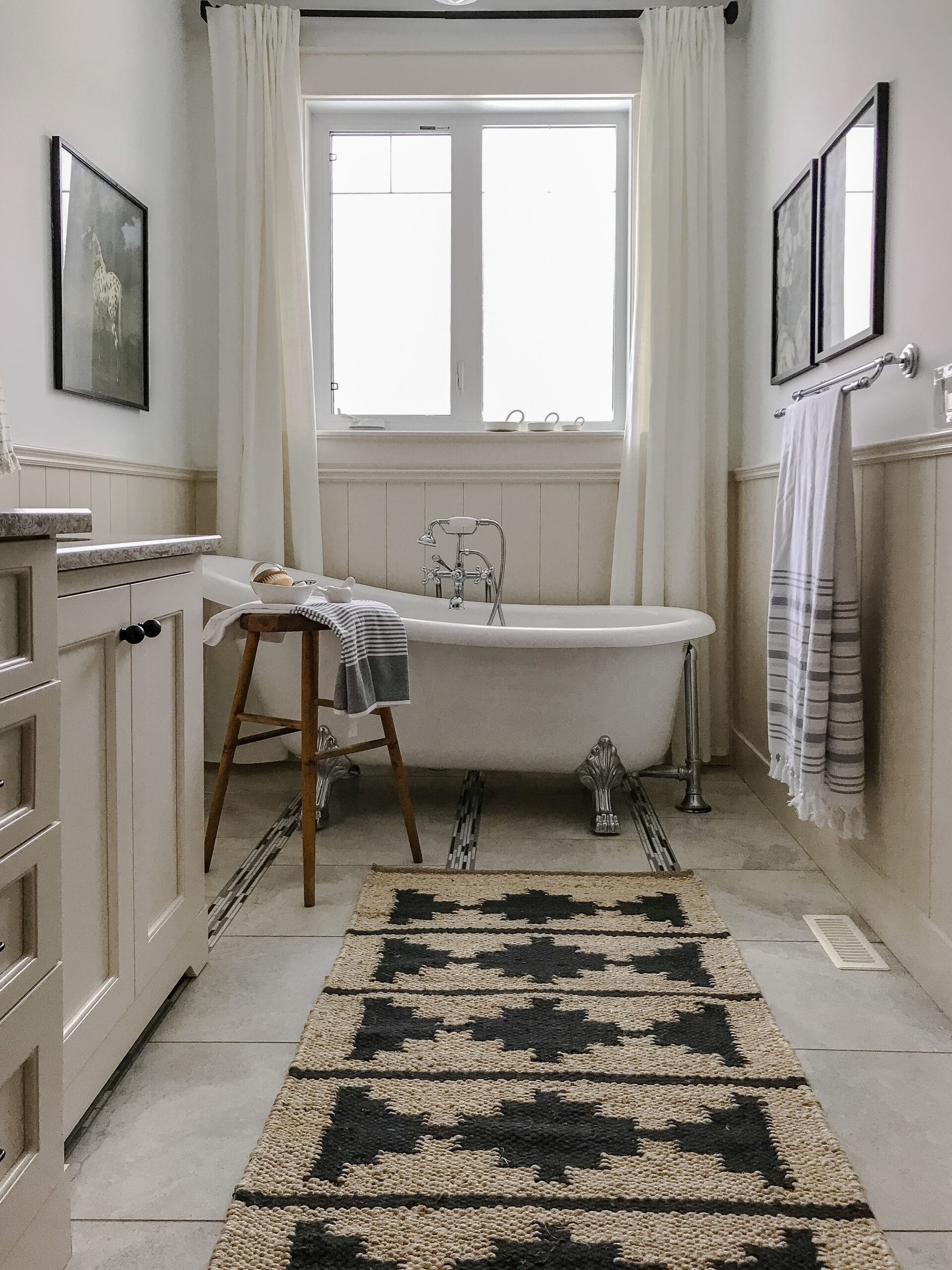 Bathroom with white walls, beige wainscotting, clawfoot tub under a window with white curtains, art on the walls, vintage wood stool, beige cabinets on the left with black hardware