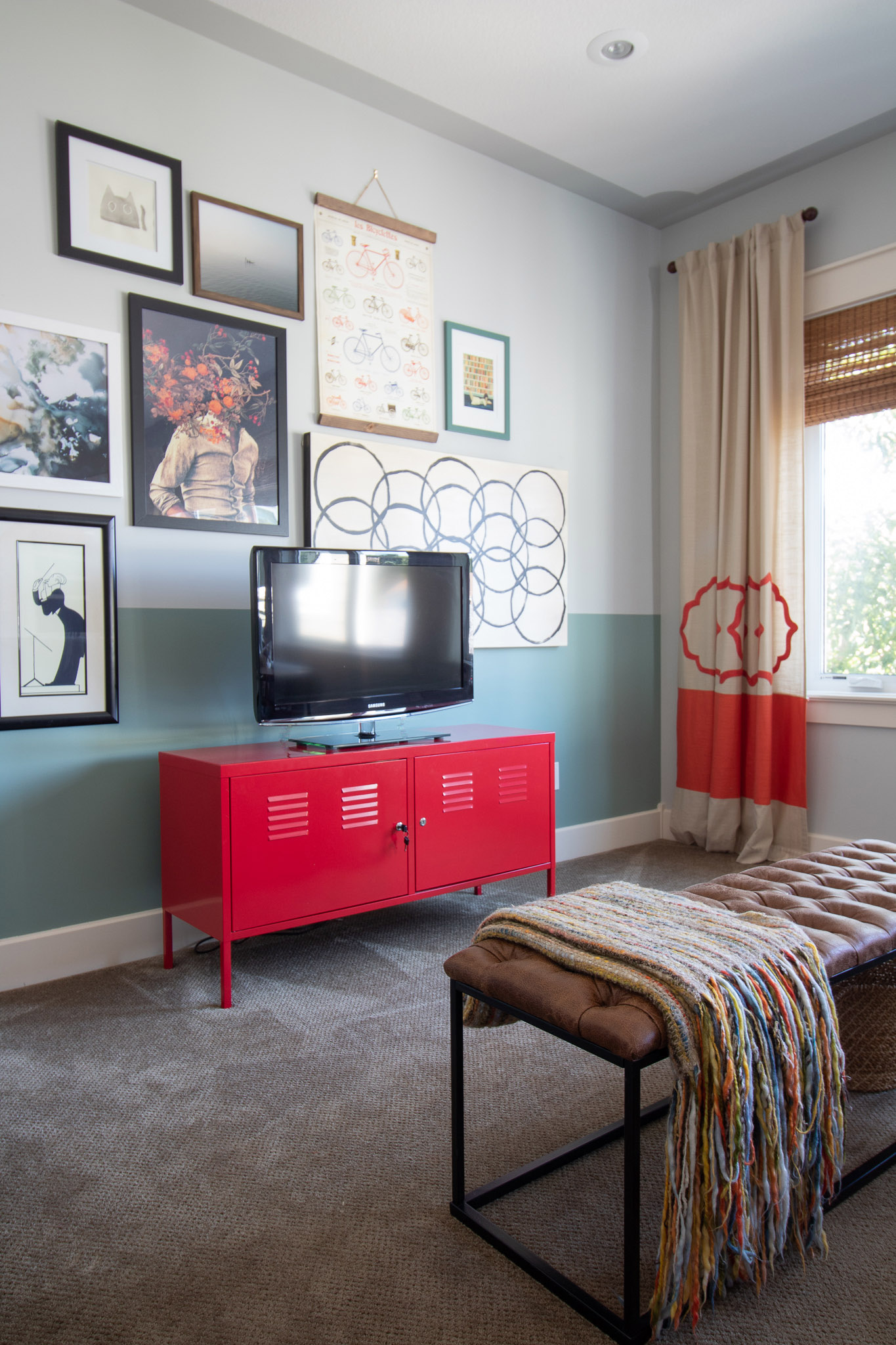 Room with a TV on a red TV stand and a green colour blocked wall with a gallery wall of art surrounding the TV.  Red patterned curtains in the background