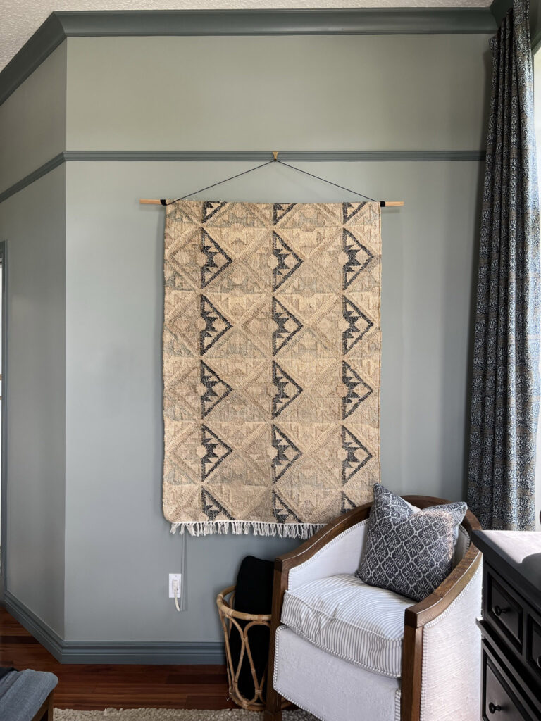 Rug hanging from a piece of wood and a string from a chair rail in a room with green walls and dark green contrast trim. Antique chair in foreground