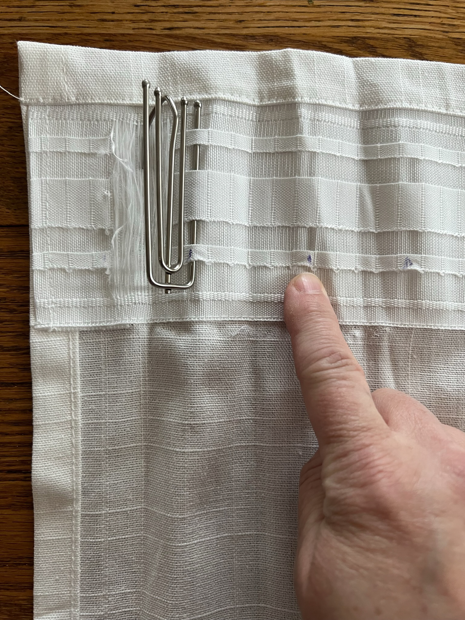 The first prong of the curtain hook has been inserted and the location of the next prong is marked with a pen, with a finger pointing to it.