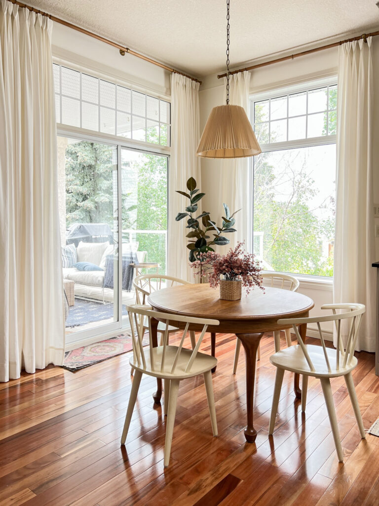 Dining nook with white walls and white DIY pinch pleat curtains, round wood table and white chairs and a pleated pendant over the table, plant in the background and pink flowers on the table.