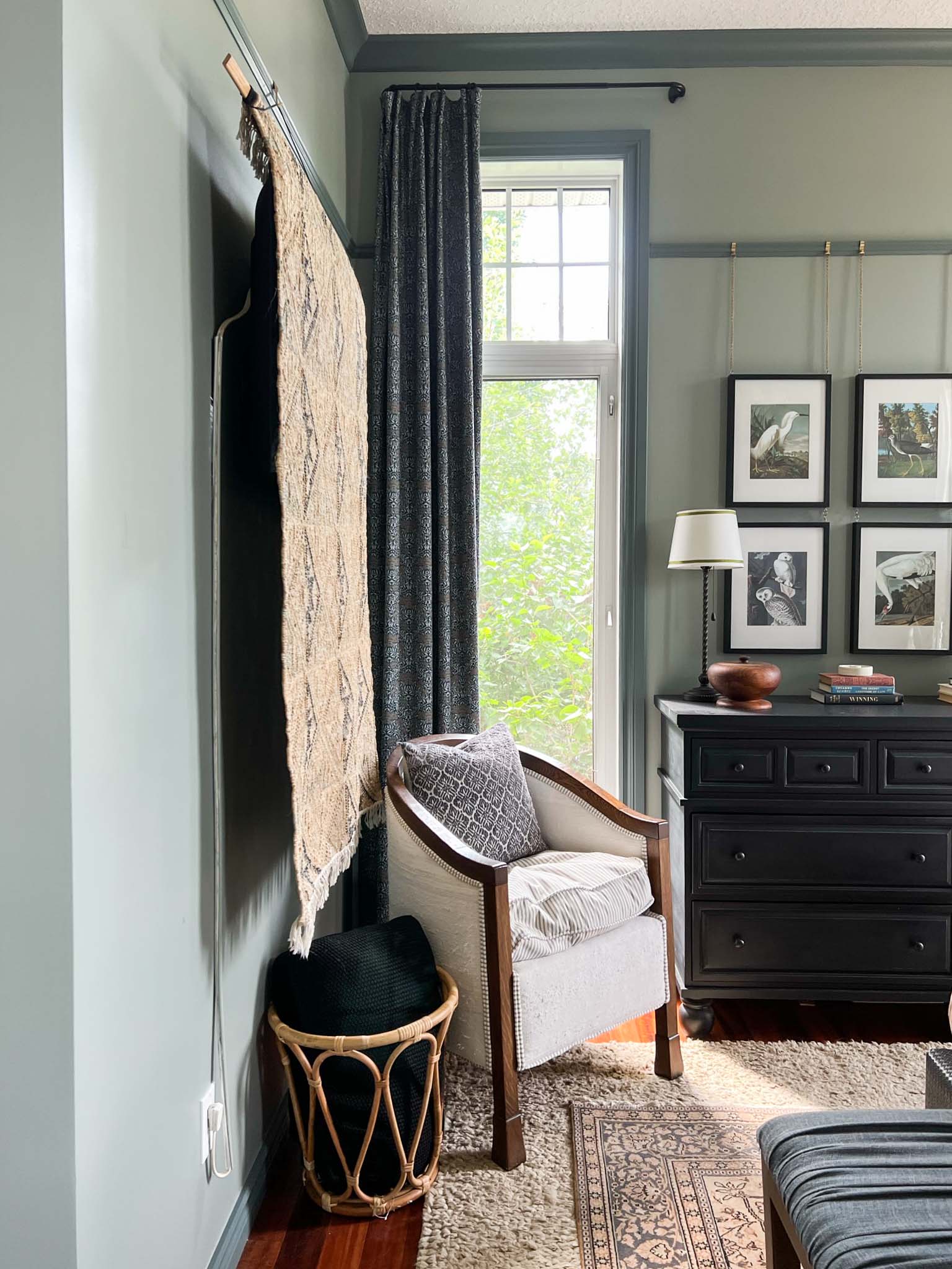 Viewing the rug hanging from the side where you can see the TV slightly.  Also can see a vintage layered rug, antique chair, basket with pillows, black dresser, long narrow window with floral curtains, green walls with dark green contrast trim.