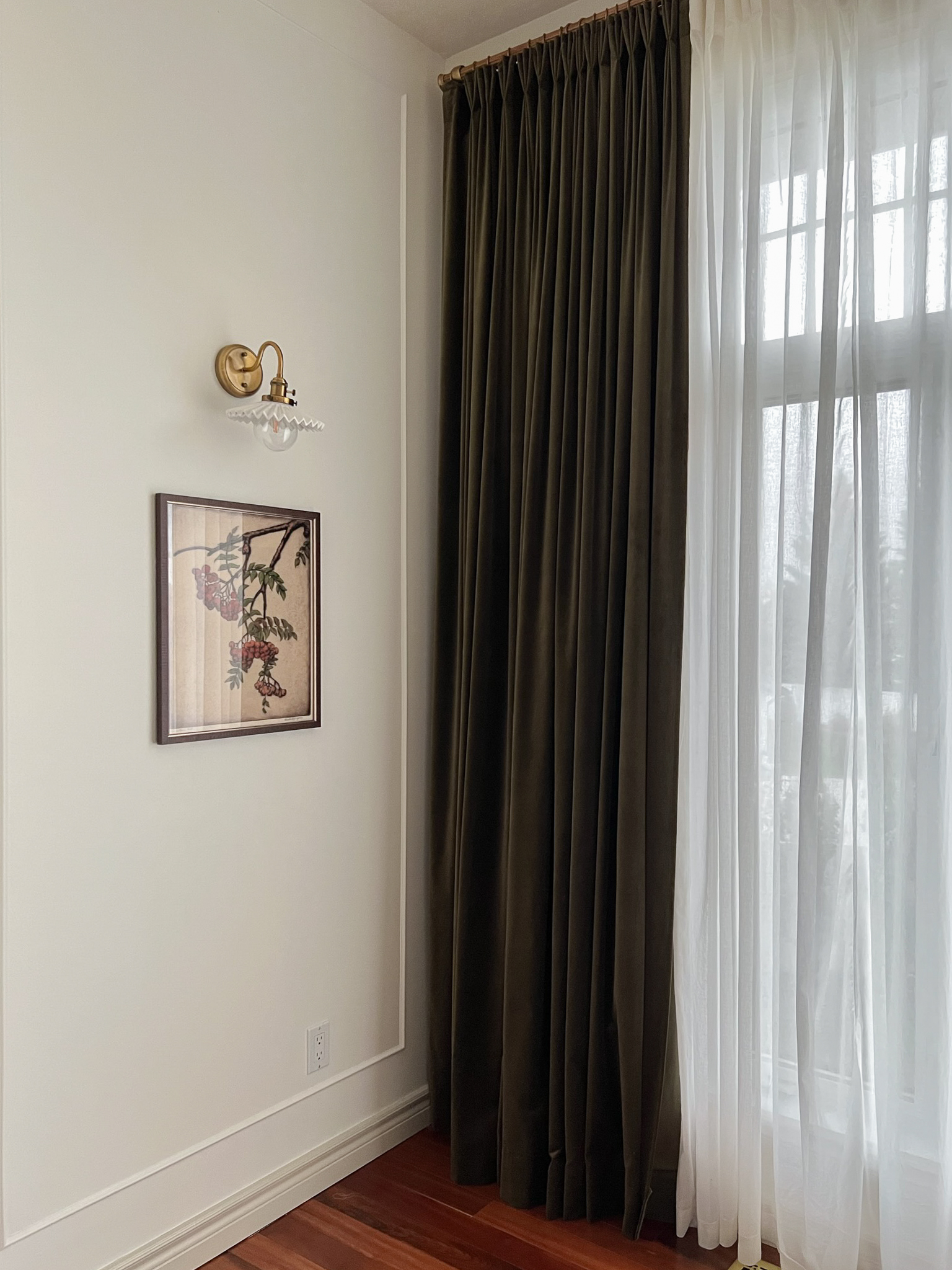 Green velvet curtains on one side of a window with DIY Pinch pleats. Wall to the left has a botantial art print with a sconce above