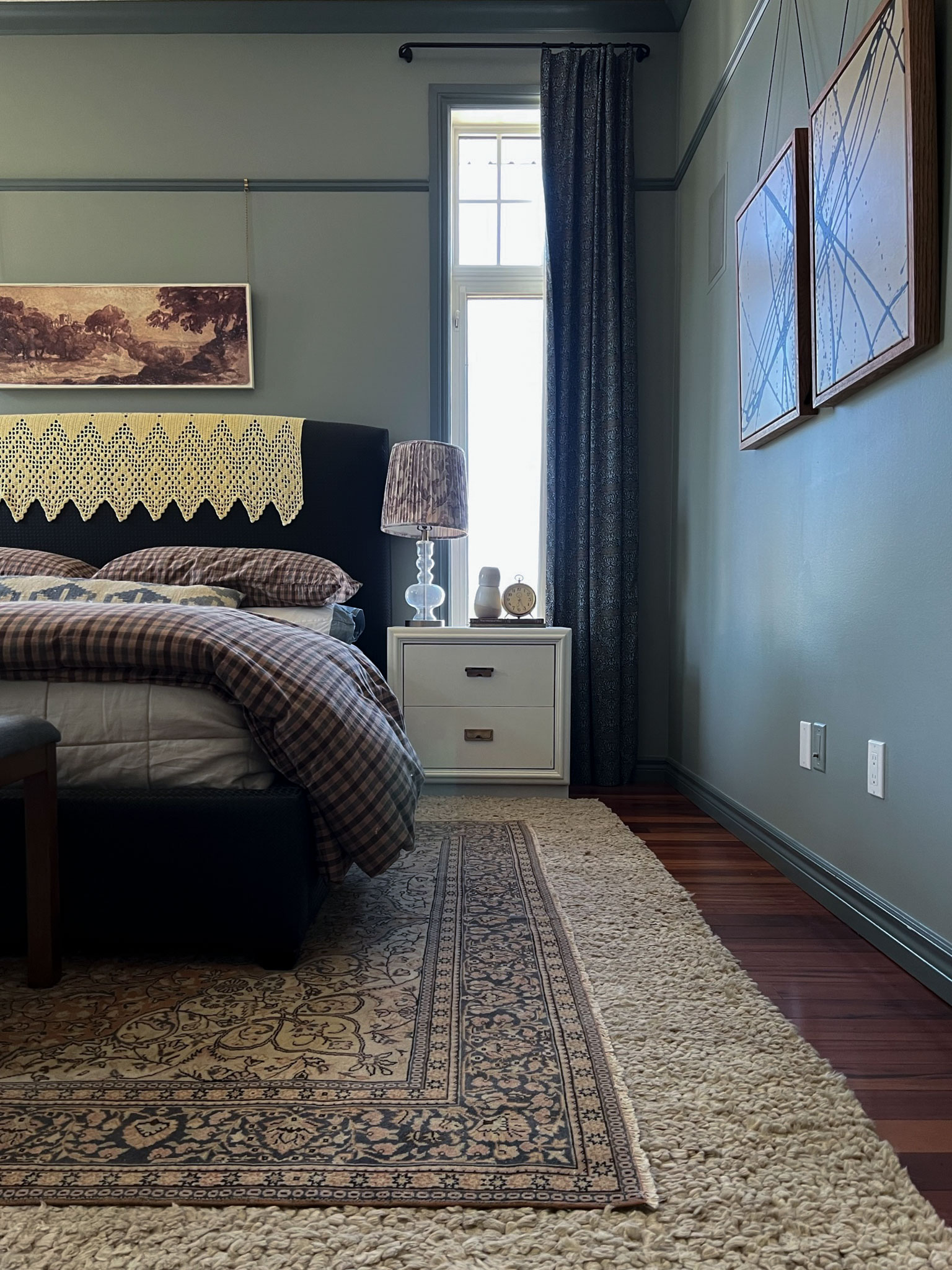 Bedroom with pale green walls and darker green trim, picture rail, beige and brown checkered bedding, black bed, bench at the end of the bed with two layered wool rugs. White nightstands.