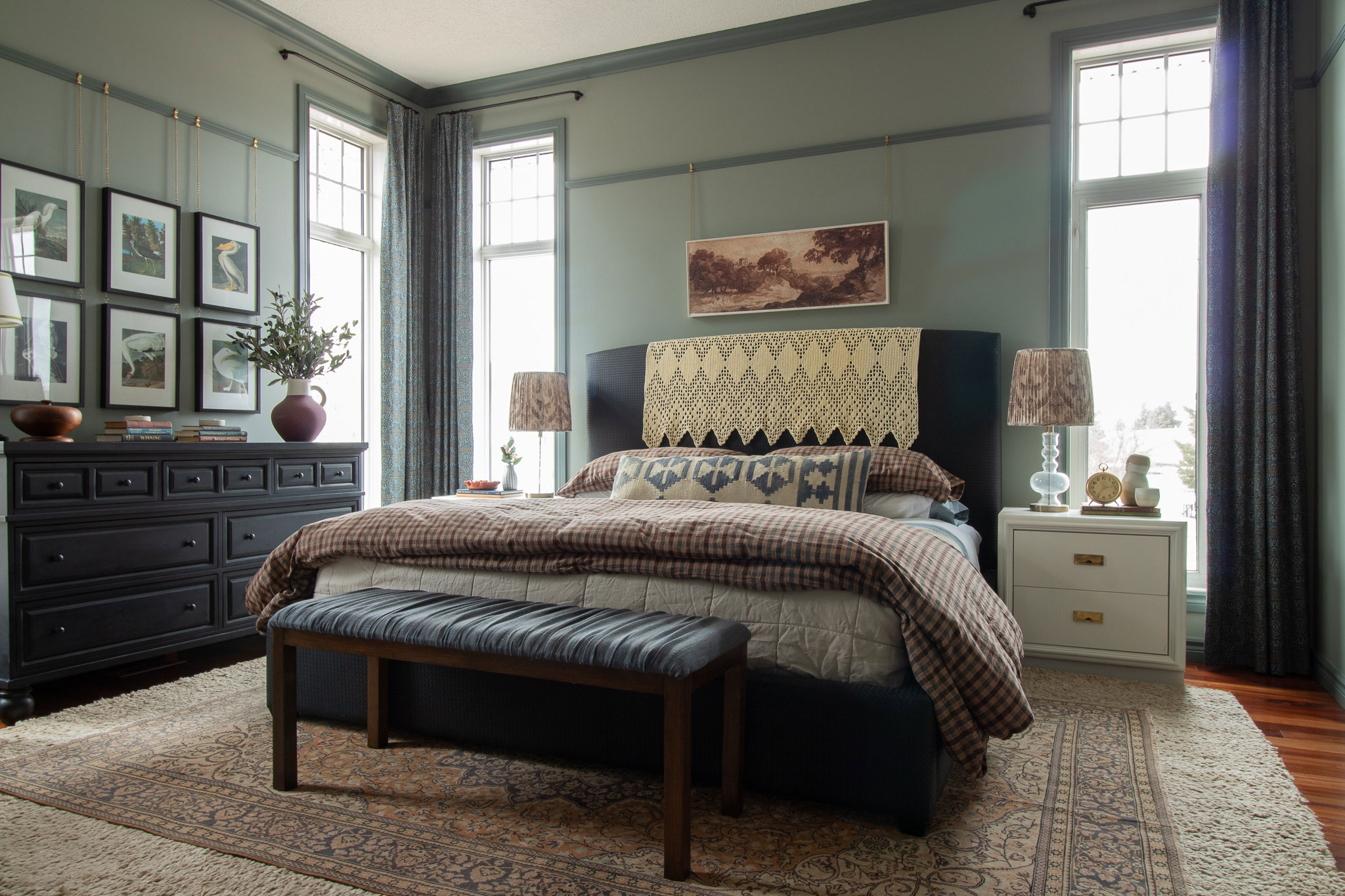 Bedroom with pale green walls and darker green trim, picture rail, beige and brown checkered bedding, black bed, bench at the end of the bed with two layered wool rugs.  White nightstands.