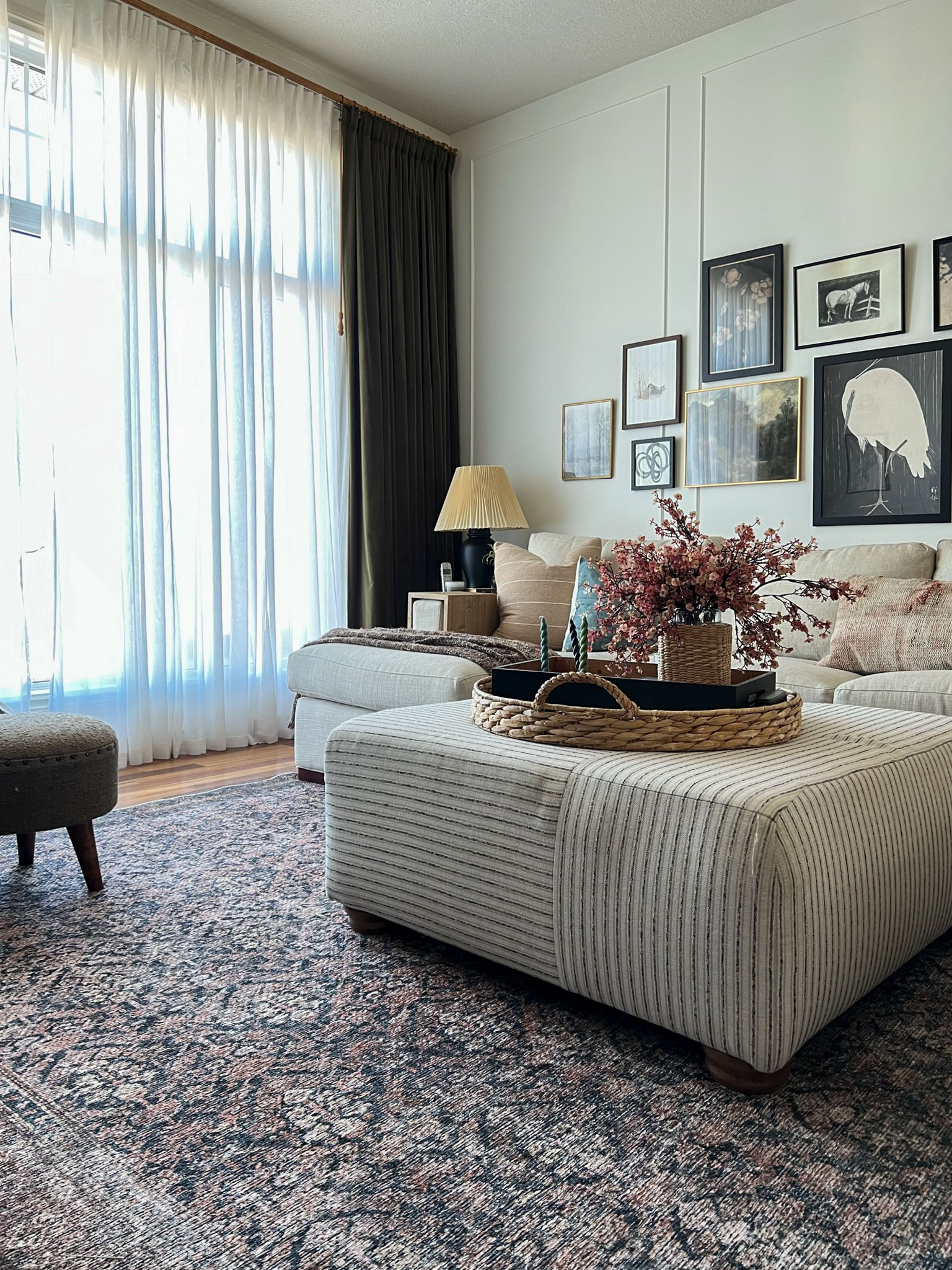 Living room with synthetic printed loloi rug, upolstered striped ottoman, gallery wall of vintage art, green velvet curtains and sheers on a large window, pink flowers