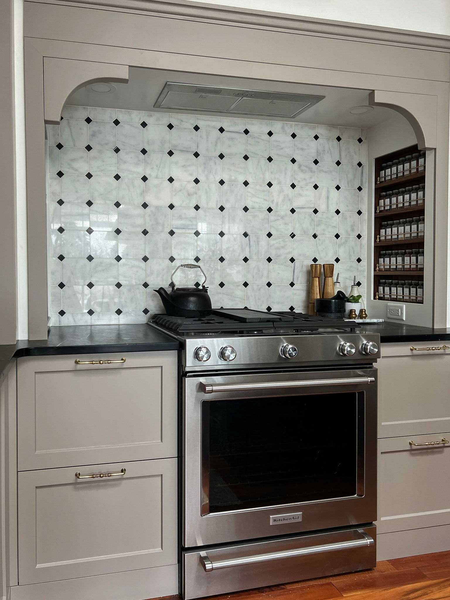 Gas ragne with beige cabinet and black soapstone countertops and octagon and dot marble tile in a stove nook with a built in DIY walnut and brass spice rack on one side.