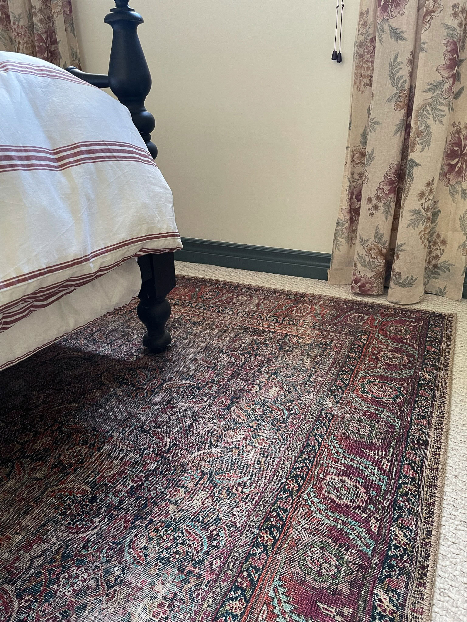 rug under a black antique bed with floral curtains and pink striped duvet, green contrast trim with white walls