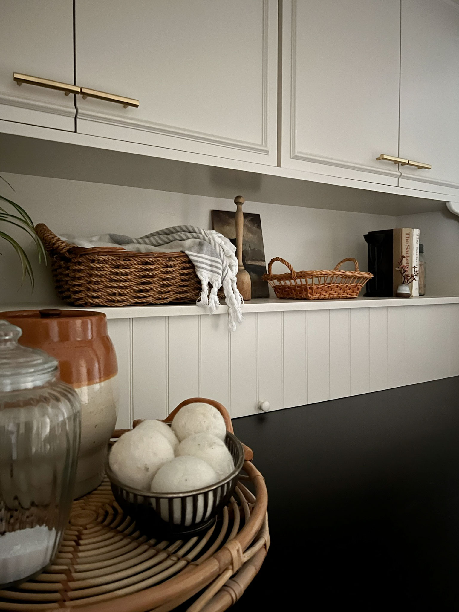 Accessories sitting on a shelf like baskets, jars and vintage paintings.  Beige shelf and cabinets above, black counters