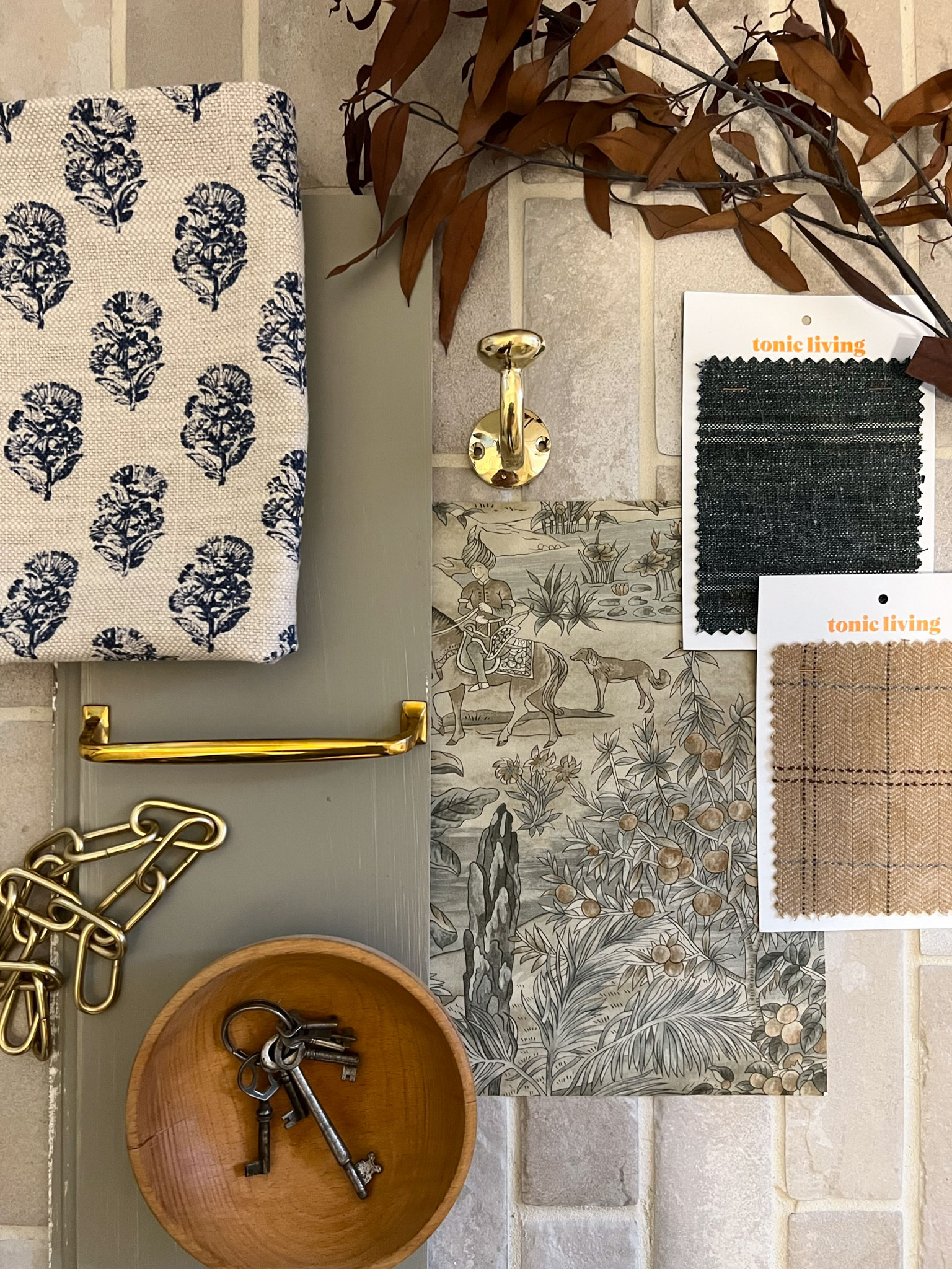 wallpaper sample, fabric samples, brass hardware and a wood bowl and brown dried leaves laying on a tile floor flatlay
