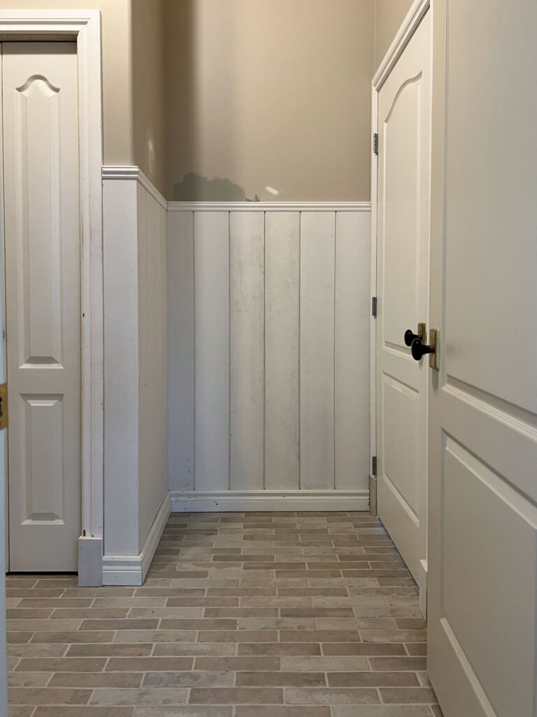 room with white unpainted shiplap wainscotting, beige brick style floors, brown walls