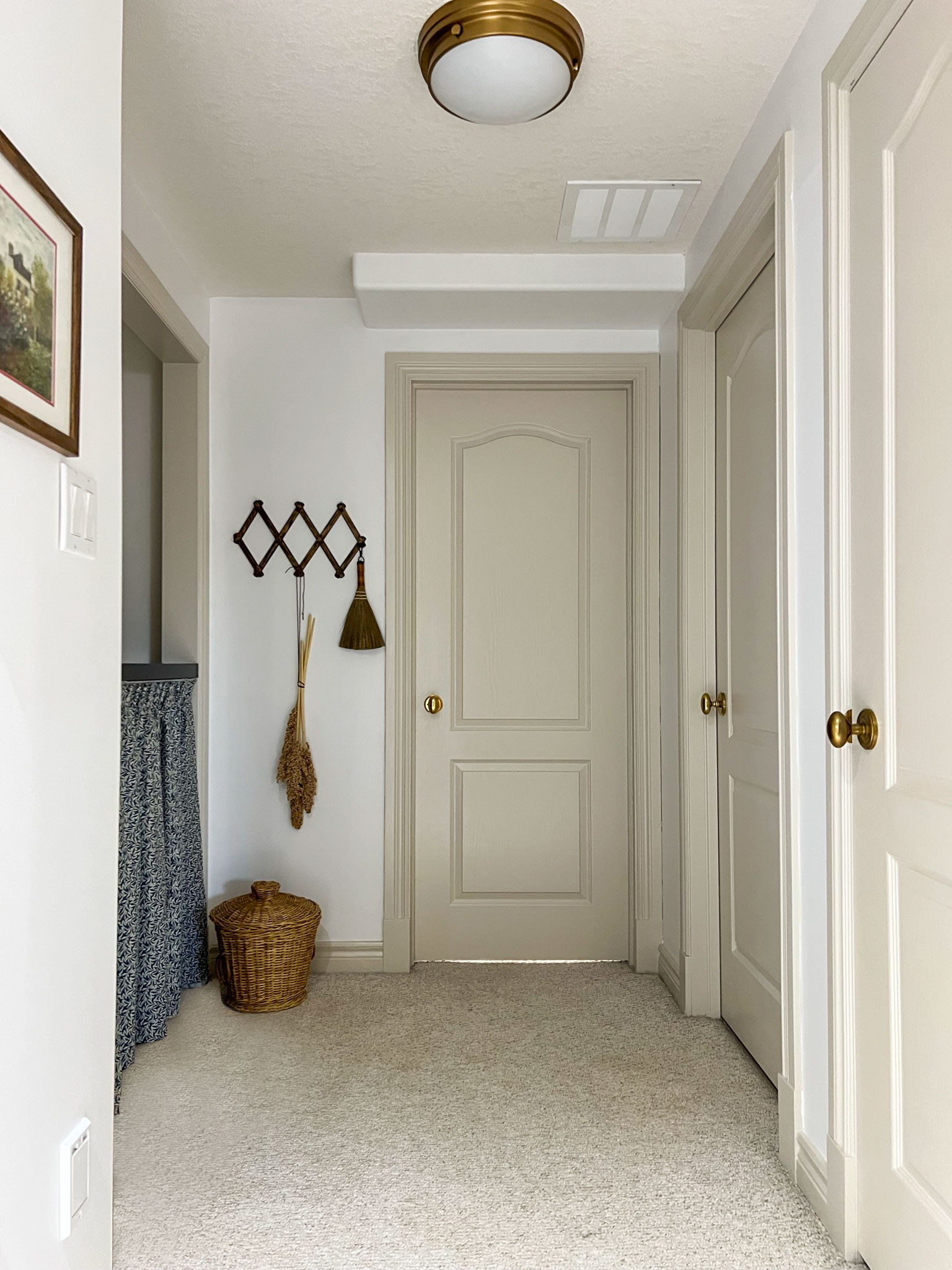 hallway with beige contrast trim and doors and white walls, accordian style hanger on wall with a broom hanging and basket on the floor, laundry closet with countertop curtain on the left