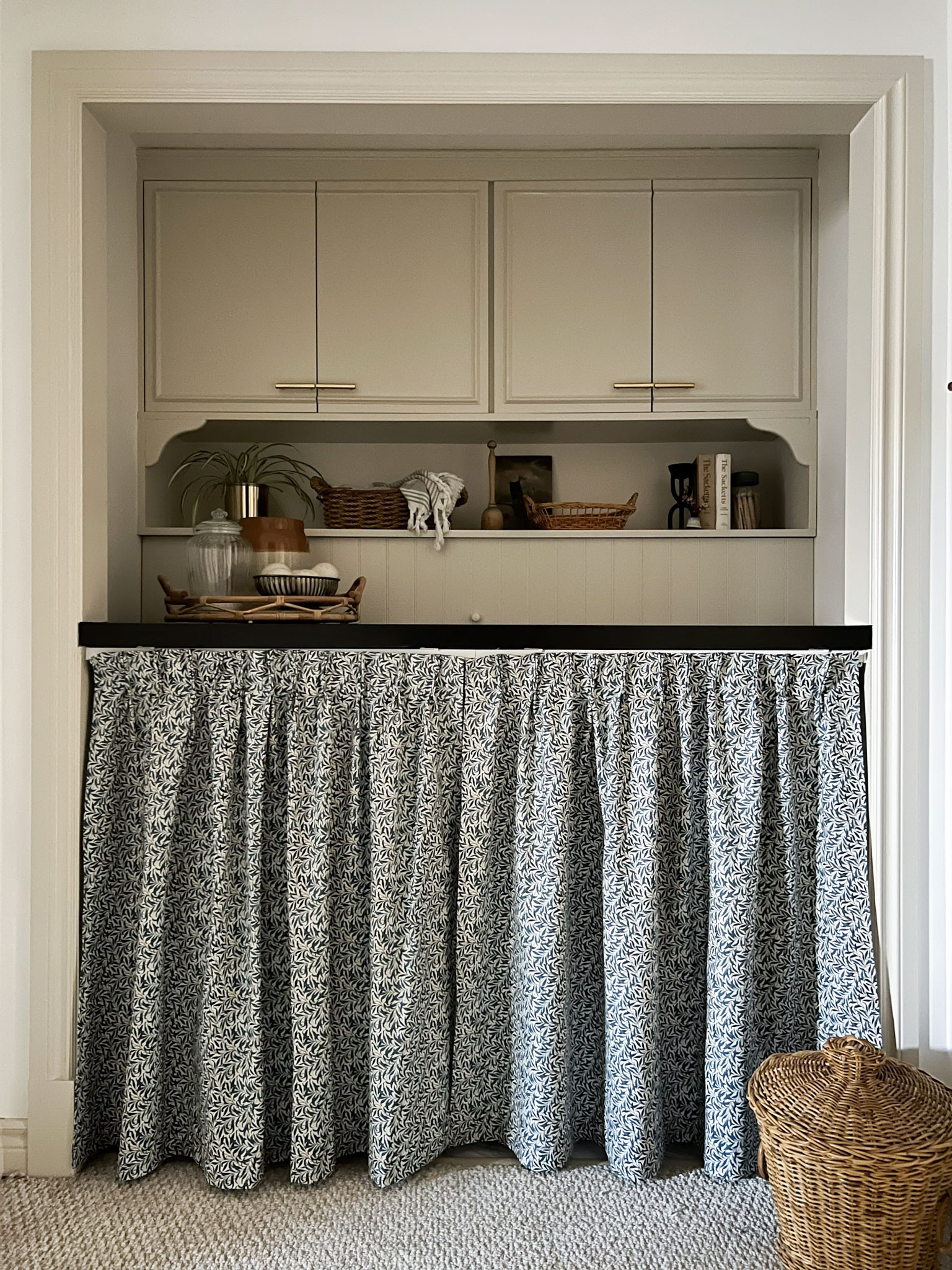 closet opening with beige cabinets in edgcomb gray, shelf below styled iwth various items, black countertop, and a william morris curtain skirt below the counter. Basket on the floor