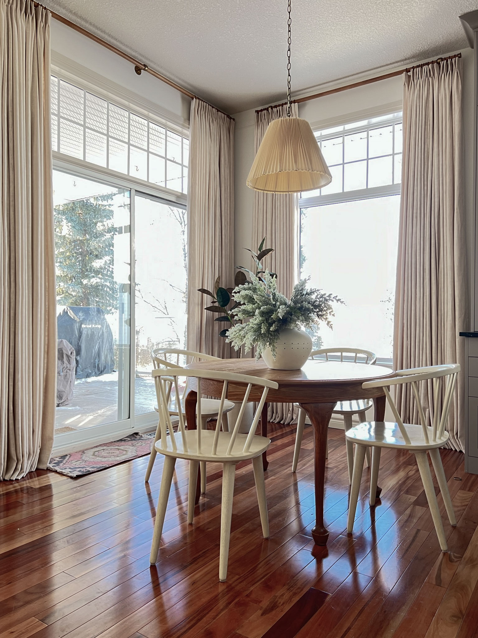 Dining nook with wood table and white chairs, pleated pendant, beige linen curtains, large vase with greenery on table