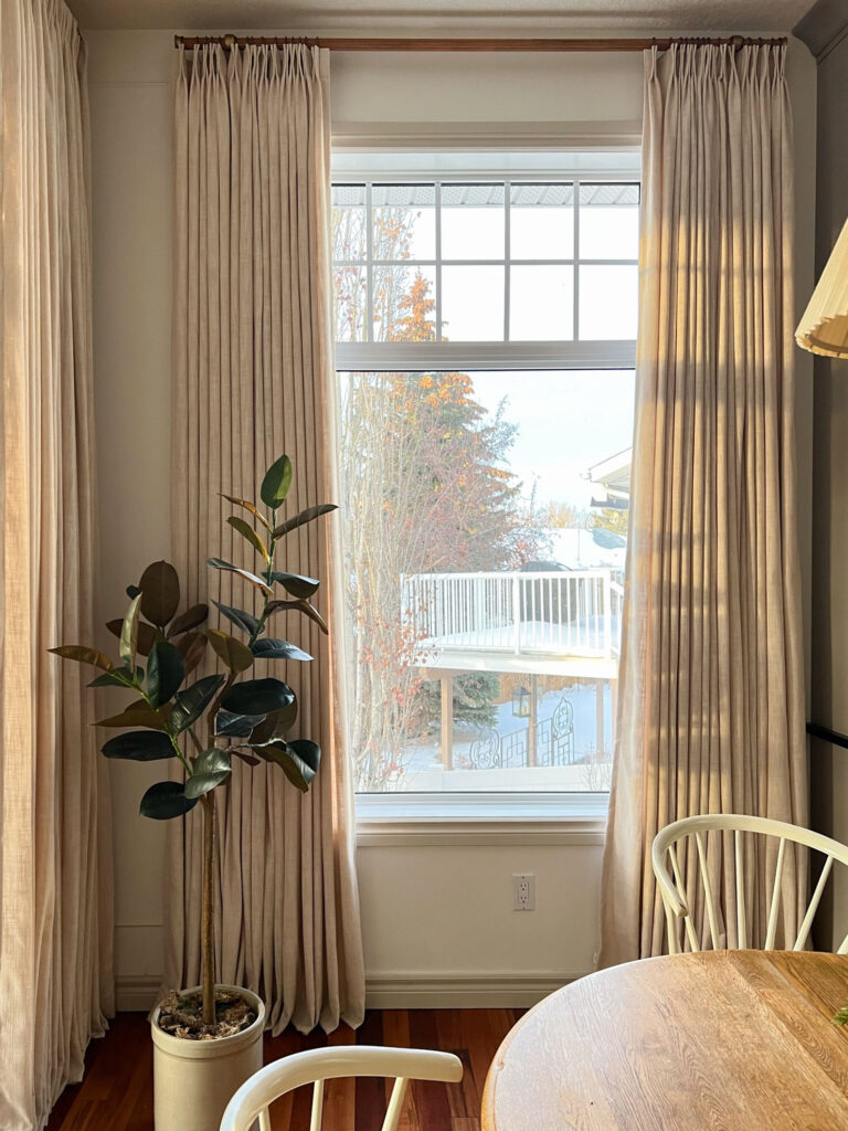 Window with very full beige curtains, white walls and beige trim, plan in a crock in front