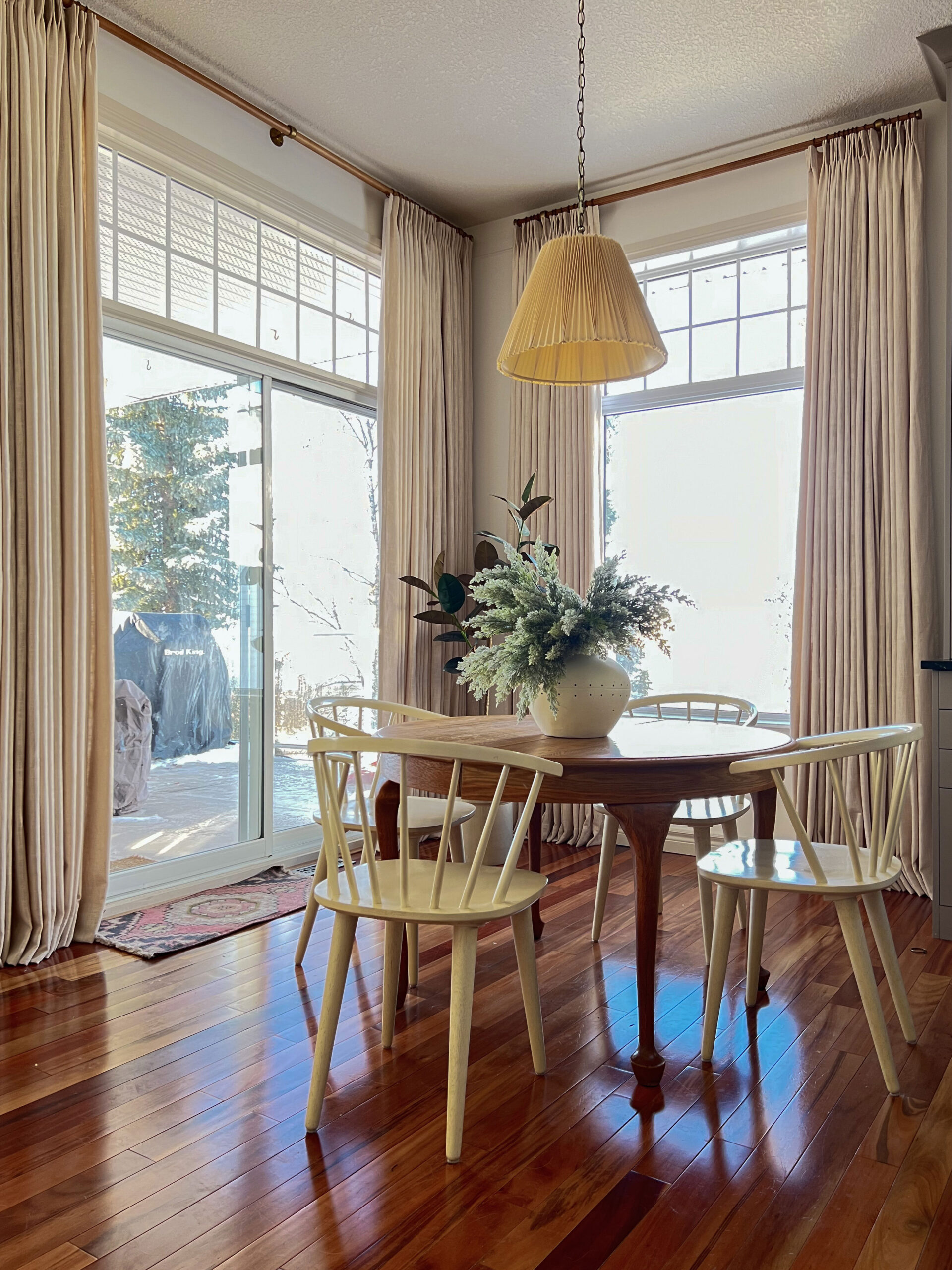 Dining nook with wood table and white chairs, pleated pendant, beige linen curtains, large vase with greenery on table