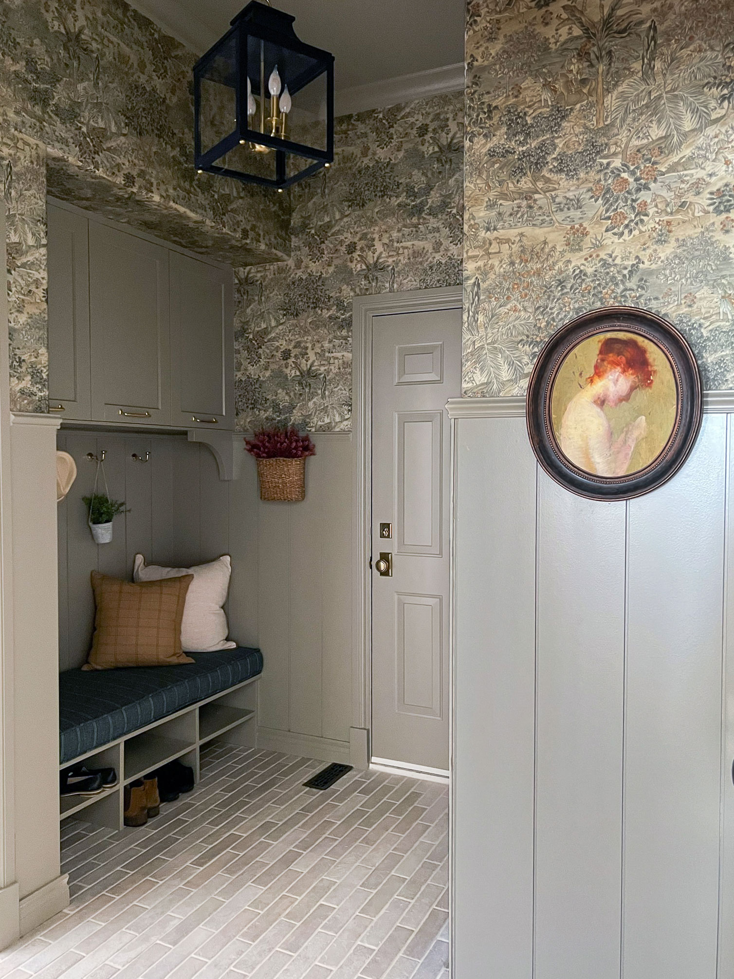 mudroom with built in bench seat, grey/green walls, beige tile brick floor, scenic wallpaper and bench cushion with pillows