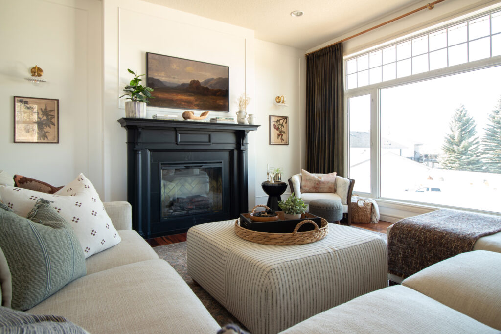 Living room with black painted mantle and black marble, upholstered ottoman, large window with green velvet curtains and wood and brass rod