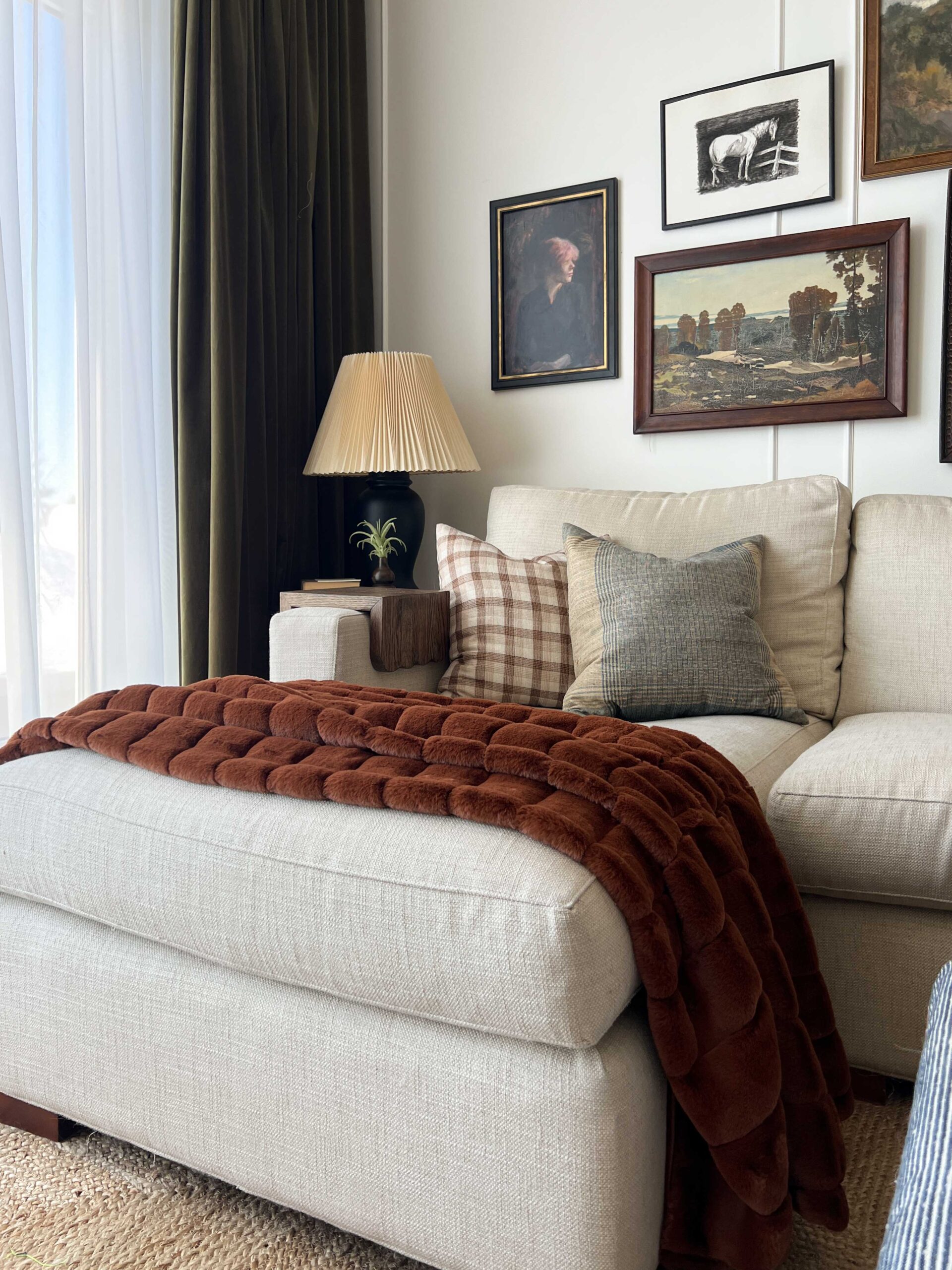corner of a sofa with a chaise and a fuzzy red blanket thrown over, pillows, green curtains and vintage art