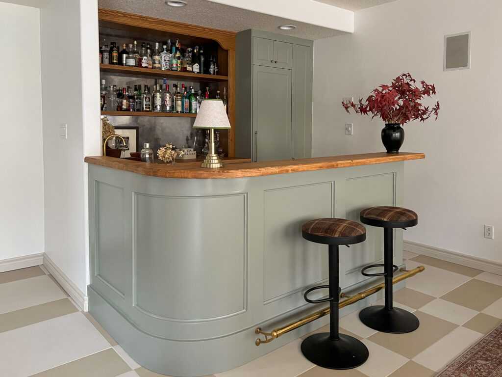 DIY bar build with green curved bar front, black and plaid stools, and a wood mirrored back bar
