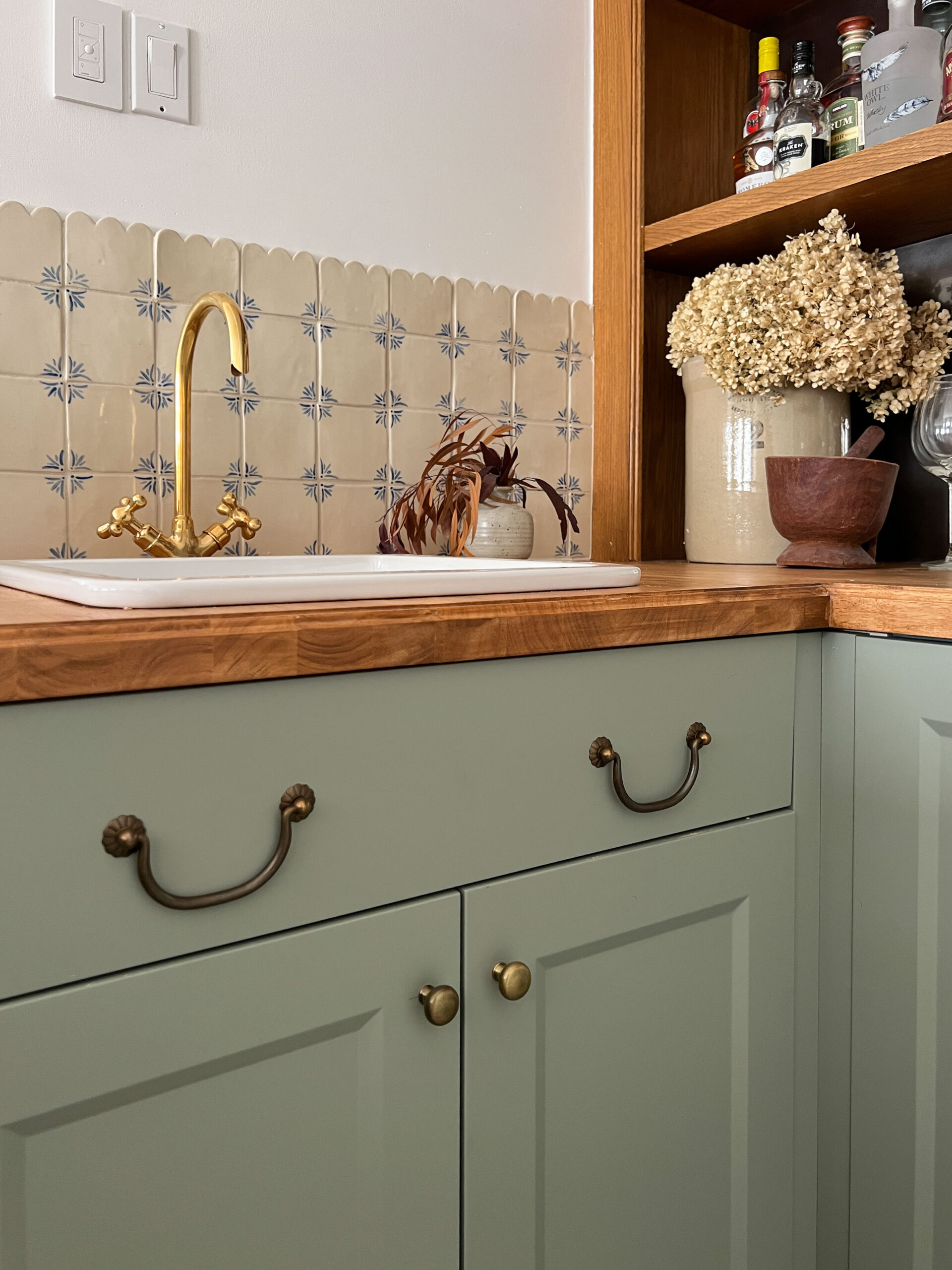 green cabinets, butcherblock counter, and delft style handmade tile with a white ceramic overmount sink and brass faucet