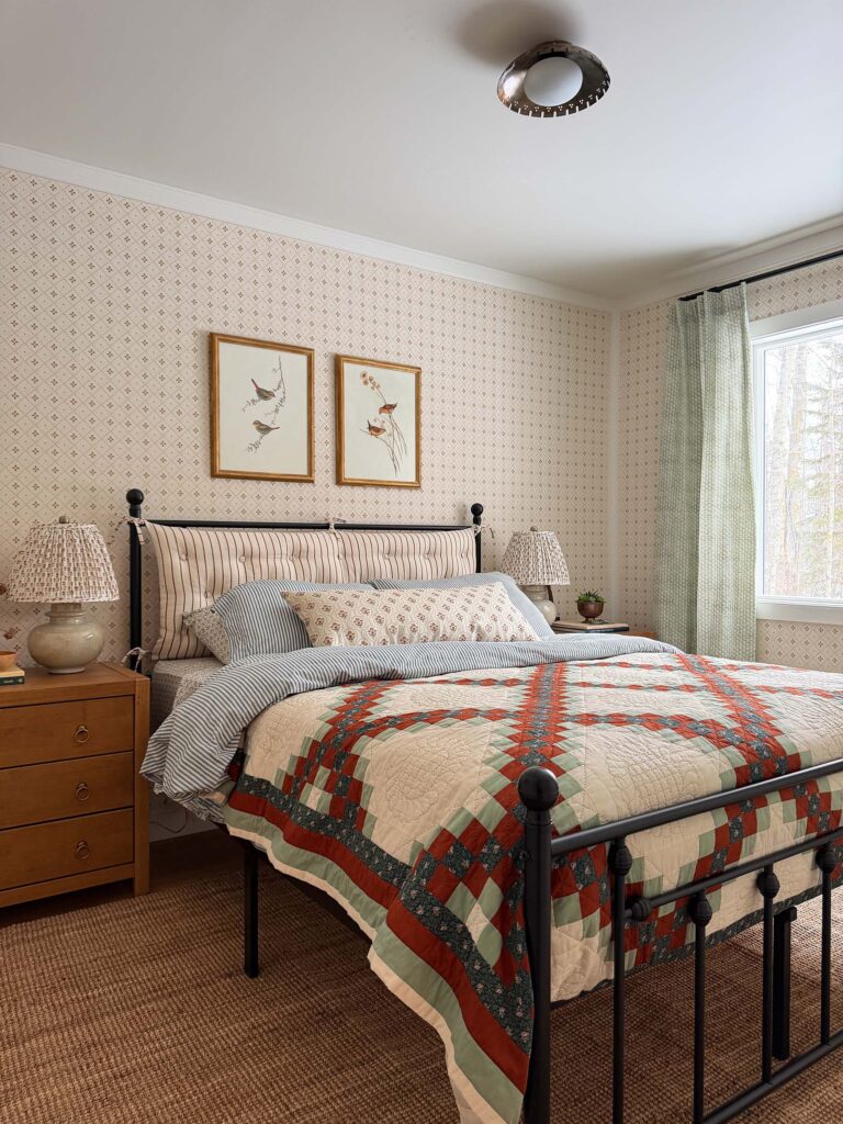 cottage style bedroom with a geometric floral neutral wallpaper, vintage quilt, patterned bedding and jute rug, wood nightstands, and an iron bed