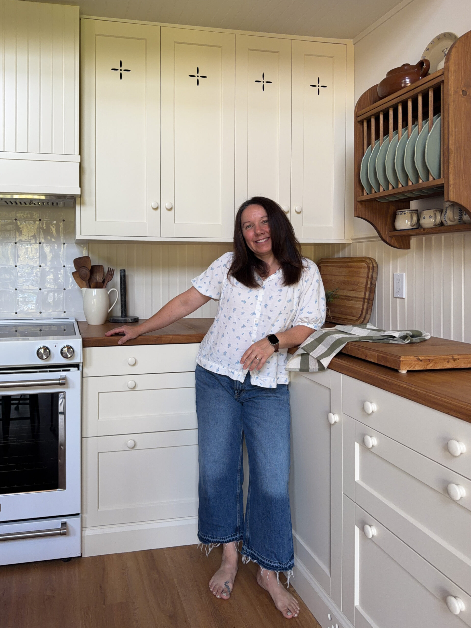 Ikea kitchen with cream coloured cabinets, butcherblock counters, antique table, freestanding wood pantry, plate rack, woman standing in kitchen